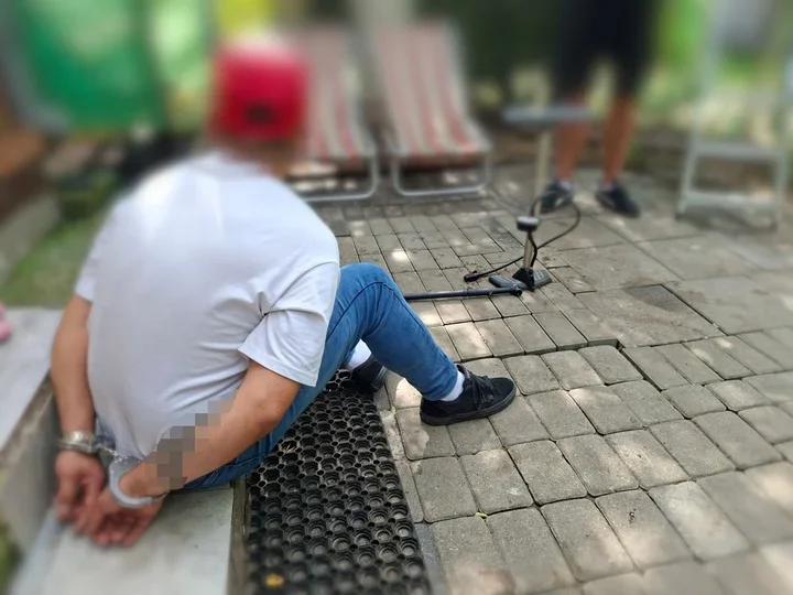 Mężczyzna w białej koszulce, niebieskich dżinsach i czerwonej czapce siedzi na chodniku z rękami skrępowanymi kajdankami za plecami, obok niego leży urządzenie
