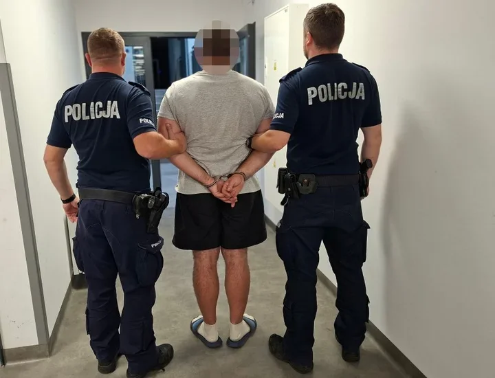 Dwóch policjantów w mundurach z napisem POLICJA prowadzi zatrzymanego mężczyznę w kajdankach korytarzem budynku.