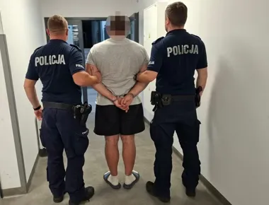 Dwóch policjantów w mundurach z napisem POLICJA prowadzi zatrzymanego mężczyznę w kajdankach korytarzem budynku.