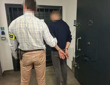 Funkcjonariusz służby więziennej prowadzi zakutego w kajdanki mężczyznę w kierunku celi.