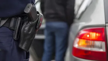 Fragment munduru policjanta z kaburą i pistoletem przy tylnym zderzaku samochodu osobowego.