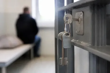 Klucz w zamku celi więziennej, w głębi niewyraźna sylwetka siedzącego mężczyzny.