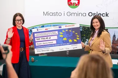 Dwie kobiety trzymają duży czek na dofinansowanie termomodernizacji szkoły w Osiecku podczas uroczystości w sali konferencyjnej.