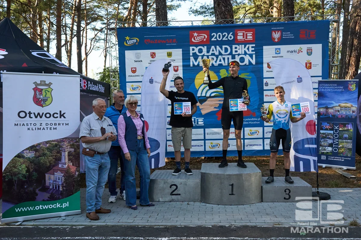 Trzech mężczyzn stoi na podium podczas dekoracji laureatów Poland Bike Marathon w Otwocku, a obok nich stoją trzy osoby.
